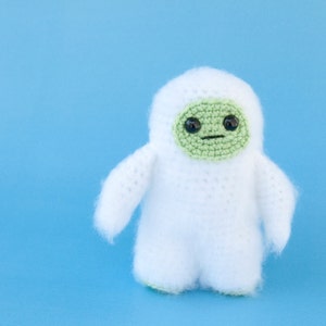 May include: A handmade, white crochet doll with a green face and black eyes. The doll has a small, round body, arms, and legs. The face is a lighter green color and has a simple, stitched mouth. The doll is set against a solid blue background.