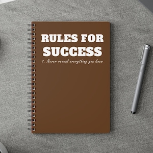 Pode incluir: Um caderno espiral marrom com o texto "RULES FOR SUCCESS" em branco, juntamente com a regra "1. Never reveal everything you know." Uma caneta prateada está ao lado do caderno. O caderno está sobre uma superfície cinza.
