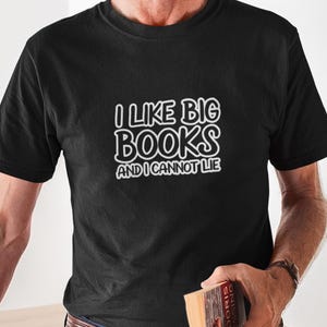 May include: Black t-shirt with the white text "I LIKE BIG BOOKS AND I CANNOT LIE." The person is holding a book with a brown cover. The shirt has short sleeves and a crew neck.