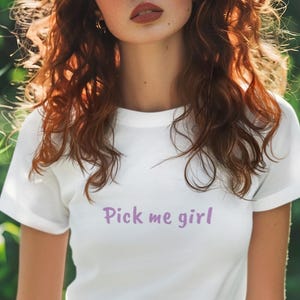 Puede incluir: Una camiseta blanca con el texto "Pick me girl" impreso en morado en la parte delantera.
