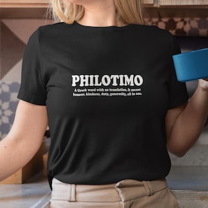 Puede incluir: Camiseta negra con la palabra "PHILOTIMO" en blanco y su definición. La definición indica que es una palabra griega que significa honor, amabilidad, deber y generosidad.