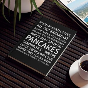 May include: A black notebook with white text listing breakfast and lunch menu items, including pancakes, sandwiches, and coffee. The notebook is on a dark wooden surface, with a coffee cup and a magazine visible.