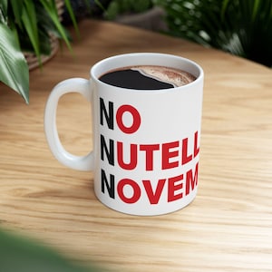 Może przedstawiać: Biały ceramiczny kubek z napisem "NO NUTELL NOVEM" w kolorze czarnym i czerwonym. Kubek jest wypełniony ciemną kawą i stoi na drewnianej powierzchni.