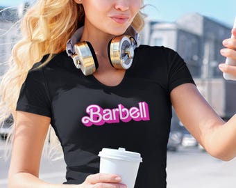 Peut inclure: T-shirt noir à col en V avec le mot "Barbell" en script rose. Le t-shirt est porté avec un casque audio doré et argenté. Une tasse en papier blanche est tenue à la main.