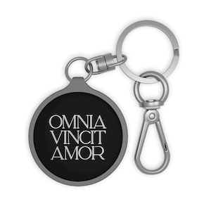 May include: A round keychain with a black face and the words "OMNIA VINCIT AMOR" in white. The keychain has a silver-colored metal ring and clasp for attaching to keys or a bag.