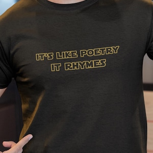 May include: Black t-shirt with the text "IT'S LIKE POETRY IT RHYMES" in a gold-coloured font. The shirt is a solid colour and has a crew neck. The image shows the upper torso of a person wearing the shirt.
