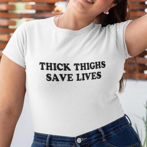 May include: A white t-shirt featuring the black text "THICK THIGHS SAVE LIVES" in a bold font. The shirt is worn with blue denim jeans. The image is a medium shot, showing the upper body.