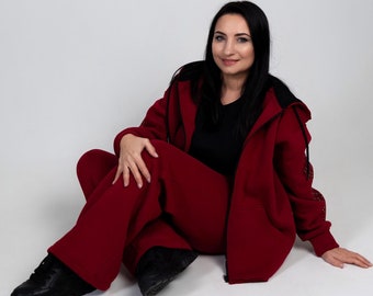 Op de afbeelding: Een vrouw in een rood hoodie-sweatshirt en bijpassende broek zit op een witte achtergrond. Het sweatshirt heeft een zwarte rits en een zwart gaaspaneel op de mouw.