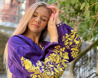 Puede incluir: Una blusa morada con mangas largas y bordados florales amarillos. La blusa tiene cuello en V y un corte holgado.
