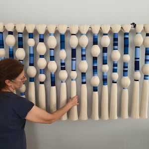 May include: A wall hanging made of white yarn with blue and white striped accents. The yarn is woven into a series of hanging strands with blue and white pom-poms.