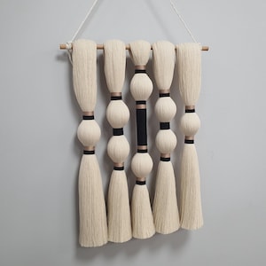Op de afbeelding: Een wandhanger gemaakt van wit garen met zwarte en gouden accenten. Het garen is geweven in een reeks kwastjes met ronde ballen ertussen.