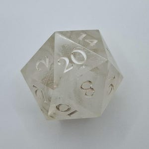 May include: A clear, translucent twenty-sided die with white numbers. The die is made of a material that resembles ice or crystal.