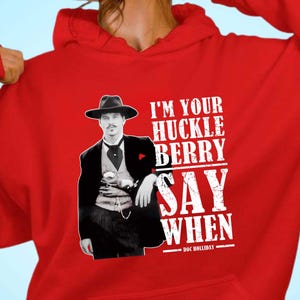 Puede incluir: Sudadera con capucha roja con un gráfico de un hombre con traje y sombrero negros, con el texto "I'M YOUR HUCKLE BERRY SAY WHEN - DOC HOLLIDAY" en blanco. La sudadera tiene una capucha con cordón y un bolsillo de canguro.