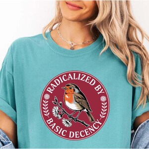 Op de afbeelding: Turquoise T-shirt met een cirkelvormige afbeelding van een roodborstje op een bloeiende tak. De afbeelding is omlijst door een bordeauxrode rand met de witte tekst "RADICALIZED BY BASIC DECENCY". Het T-shirt heeft korte mouwen.