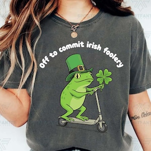 May include: Dark gray t-shirt featuring a green frog wearing a leprechaun hat and holding a four-leaf clover, riding a scooter. The text "off to commit irish foolery" is arched above the frog. A fun St. Patrick's Day graphic.