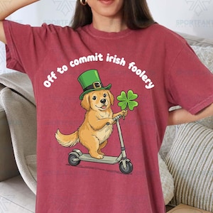 以下が含まれることがあります： 緑色の帽子をかぶり、四つ葉のクローバーを持ったゴールデンレトリバーがスクーターに乗っているイラストが描かれた、マルーン色のTシャツ。「off to commit irish foolery」の文字が犬の上にアーチ状に書かれています。