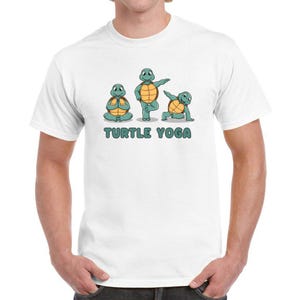 Op de afbeelding: Wit T-shirt met een afbeelding van drie cartoon schildpadden die yoga-houdingen aannemen. De tekst "TURTLE YOGA" is onder de schildpadden afgedrukt.