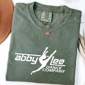 May include: An olive green t-shirt featuring the white text "abby lee DANCE COMPANY". A gold necklace with a pendant is placed on the folded shirt. The shirt has a label at the neckline.