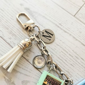 Op de afbeelding: Een zilverkleurige sleutelhanger met een witte kunstlederen kwast, een ronde bedel met de letter M, een kleine ronde strass-bedel en een miniatuur boekbedel met de tekst F. SCOTT FITZGERALD TENDER IS THE NIGHT.