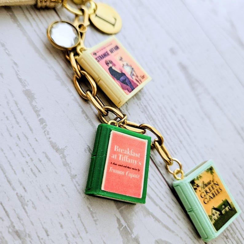Op de afbeelding: Een goudkleurige metalen bedelarmband met drie miniatuurboekbedels met de titels Strange Affair, Breakfast at Tiffany's van Truman Capote en Anne of Green Gables, samen met een ronde bedel gegraveerd met de letter L en een helder kristal.