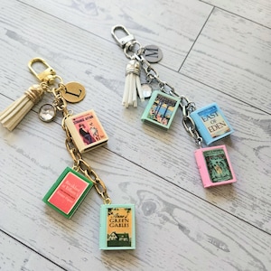 May include: Two metal keychains featuring miniature book charms. The gold-tone keychain includes charms for Breakfast at Tiffany, Anne of Green Gables, and Strange Affair. The silver-tone keychain features East of Eden and Little Women.