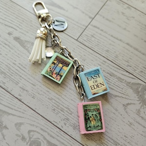 Op de afbeelding: Een zilverkleurige metalen sleutelhanger met een witte kwast, een ronde bedel met de letter M en drie miniatuurboekbedels met de titels Tender Is the Night, East of Eden en Little Women, bevestigd aan een stevige schakelketting.