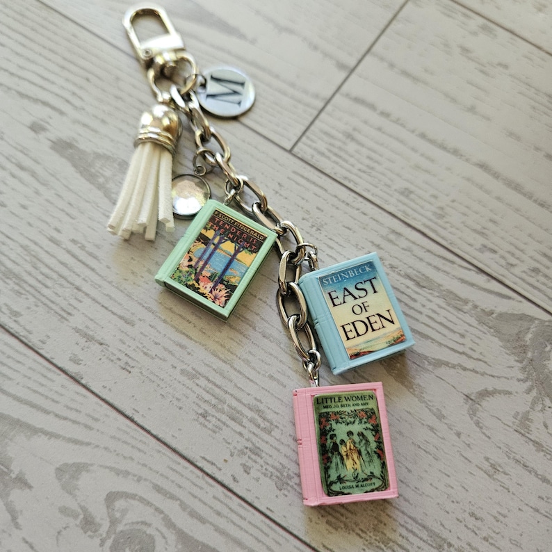 Op de afbeelding: Een zilverkleurige metalen sleutelhanger met een witte kwast, een ronde bedel met de letter M en drie miniatuurboekbedels met de titels Tender Is the Night, East of Eden en Little Women, bevestigd aan een stevige schakelketting.