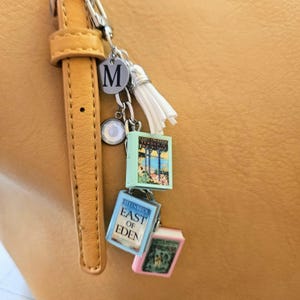 Op de afbeelding: Een zilverkleurige metalen sleutelhanger met een ronde bedel met de letter M, een witte kwast en drie miniatuurboekbedels. De boeken zijn getiteld Tender Is the Night, East of Eden en een derde boek met een roze rand en illustratie.
