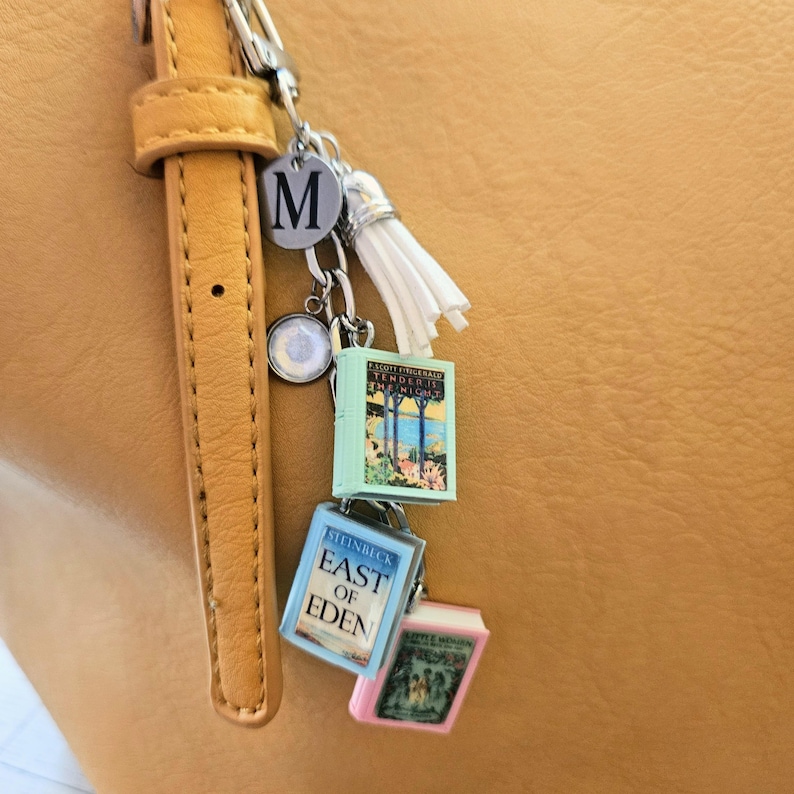 Op de afbeelding: Een zilverkleurige metalen sleutelhanger met een ronde bedel met de letter M, een witte kwast en drie miniatuurboekbedels. De boeken zijn getiteld Tender Is the Night, East of Eden en een derde boek met een roze rand en illustratie.