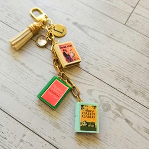 Op de afbeelding: Een goudkleurige sleutelhanger met een cr&egrave;mekleurig kwastje, een ronde bedel met de letter L en drie miniatuur boekbedels met de titels Strange Affair, Breakfast at Tiffany's en Anne of Green Gables, bevestigd aan een gouden ketting.