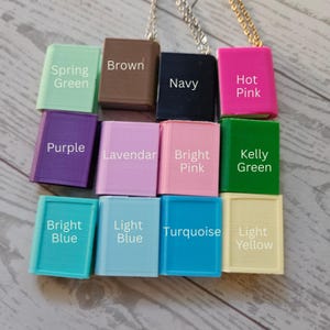 Op de afbeelding: Een verzameling van twaalf rechthoekige, boekvormige hangers aan metalen kettingen, elk gelabeld met de kleur: Spring Green, Brown, Navy, Hot Pink, Purple, Lavendar, Bright Pink, Kelly Green, Bright Blue, Light Blue, Turquoise en Light Yellow.
