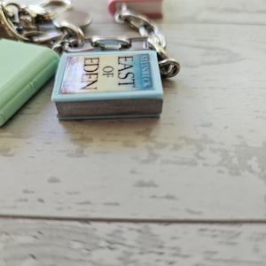 Op de afbeelding: Een zilverkleurige metalen bedelarmband met miniatuur boekbedels. Een opvallende bedel is een lichtblauw boek met de titel East of Eden van Steinbeck. Een andere bedel is een effen lichtgroen boek. De bedels zijn bevestigd met zilveren ringetjes.