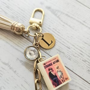 Op de afbeelding: Een goudkleurige sleutelhanger met een witte kwast, een ronde bedel gegraveerd met de letter L, een heldere gefacetteerde kristallen bedel en een miniatuurboekje getiteld Strange Affair met een vintage illustratie op de kaft.
