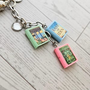Op de afbeelding: Een zilverkleurige metalen bedelarmband met drie miniatuur boekbedels. De bedels bestaan uit een lichtgroen boek getiteld Tender Is the Night, een lichtblauw boek getiteld East of Eden en een roze boek getiteld Little Women.