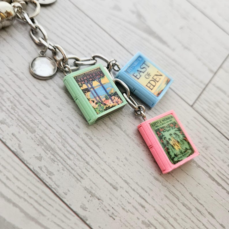 Op de afbeelding: Een zilverkleurige metalen bedelarmband met drie miniatuur boekbedels. De bedels bestaan uit een lichtgroen boek getiteld Tender Is the Night, een lichtblauw boek getiteld East of Eden en een roze boek getiteld Little Women.