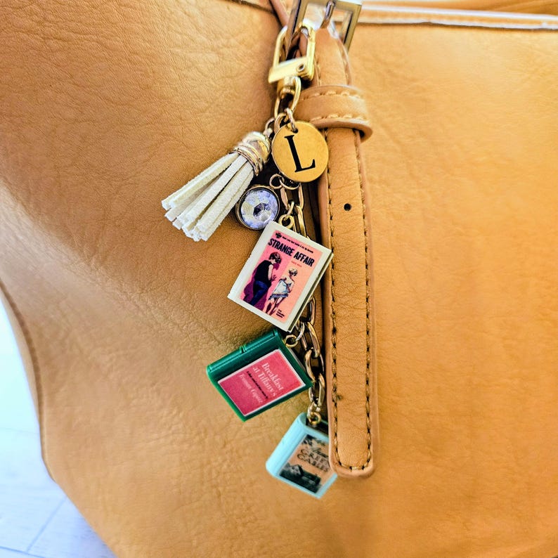 Op de afbeelding: Een goudkleurige tashanger bevestigd aan een beige leren tas. De hanger bevat een witte kwast, een ronde schijf met de letter L, een helder kristal en drie miniatuurboekhangers met de titels Strange Affair, Breakfast at Tiffany's en Green Gables.