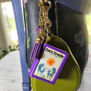 May include: A purple and gold book charm with a tassel, suspended from a gold chain. The book cover reads "Cat's Cradle" by Kurt Vonnegut, Jr., with an illustration of hands and a sun. The charm is attached to a green leather item.