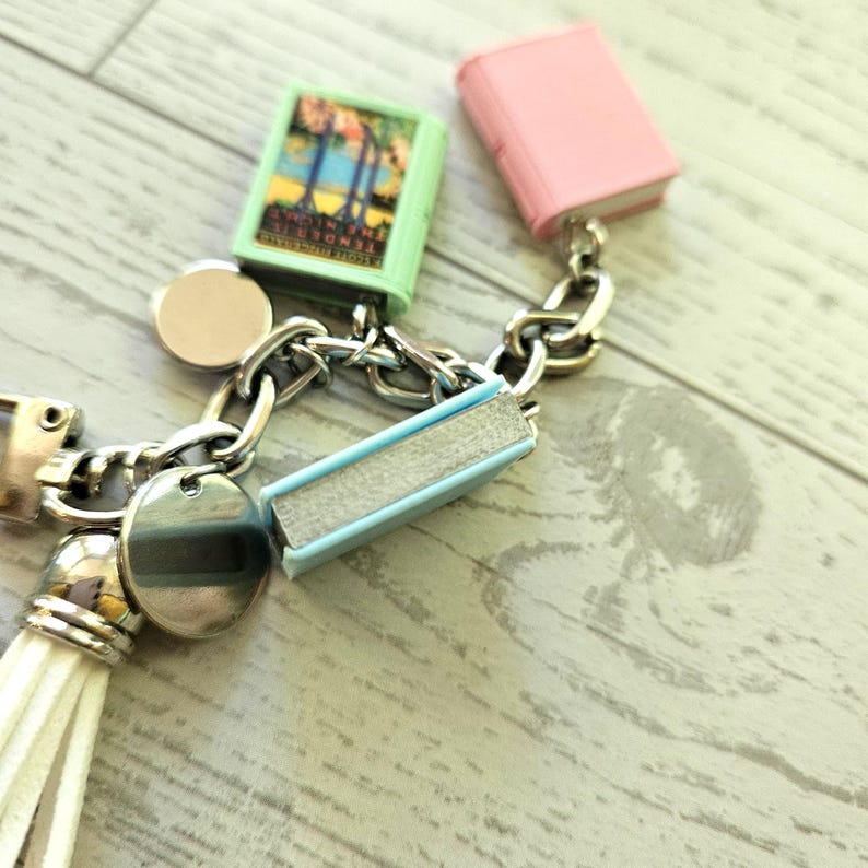Op de afbeelding: Een zilverkleurige bedelarmband met miniatuur boekbedels in lichtgroen, roze en lichtblauw, samen met twee ronde zilveren schijfjes en een witte kunstlederen kwastje. De groene boekbedel toont de tekst Tender Is The Night.