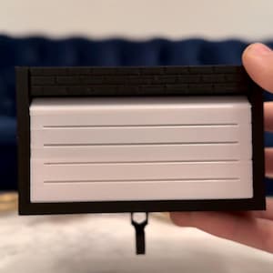 May include: A miniature garage model with a black frame and a white, slatted door. The top features a brick-like design. A small, black handle hangs from the bottom. The model is held in a person's hand.