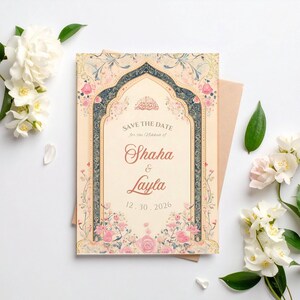 May include: A wedding invitation with a floral design. The invitation features the names "Shaha & Layla" and the date "12.30.2026". The design includes pink and blue floral accents and is set against a cream background. A cream envelope is behind the invitation.