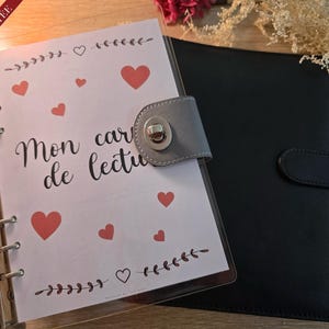 May include: A white planner with a clear cover and a black leather binder. The planner has the text "Mon carnet de lecture" and hearts printed on it. The cover has a grey snap closure.