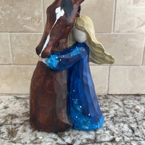 May include: A wooden sculpture of a girl hugging a horse. The girl is wearing a blue dress with white stars and has blonde hair. The horse is brown with a white stripe on its face.