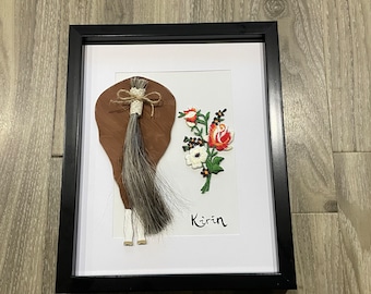 May include: Framed artwork with a horse tail and embroidered flowers. The horse tail is brown and grey, with a white base, tied with twine. The flowers are red, white, and green. The name "Kirin" is written below the flowers.