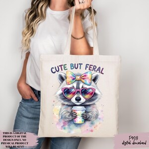May include: A cream tote bag with a raccoon illustration wearing heart-shaped sunglasses and a bow, holding a coffee cup. The text "CUTE BUT FERAL" is above the raccoon. The bag is held by a person. The text "THIS IS A DIGITAL PRODUCT OF THE DESIGN ONLY. NO PHYSICAL PRODUCT WILL BE SENT" is on the bottom left.