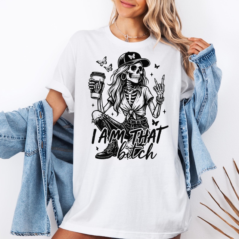 K&ouml;nnte beinhalten: Wei&szlig;es T-Shirt mit einem schwarz-wei&szlig;en Grafikdesign eines Skeletts mit Baseballkappe und Kaffeetasse. Der Text "I AM THAT bitch" steht darunter. Schmetterlinge und Sterne sind ebenfalls im Design enthalten.