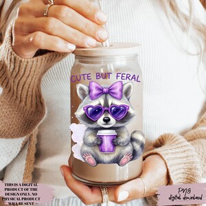 May include: A glass tumbler with a wooden lid and a straw. The tumbler features a cartoon raccoon wearing purple heart-shaped sunglasses and a bow, holding a purple cup. The text "CUTE BUT FERAL" is above the raccoon. The image also includes the text "PNG digital download".