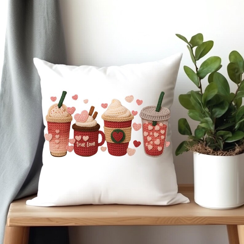May include: White square pillow featuring four crocheted coffee drinks in shades of red, pink, and beige. The pillow has a Valentine's Day theme, with heart accents and the words "True Love" on one cup. A green plant in a white pot sits to the right.