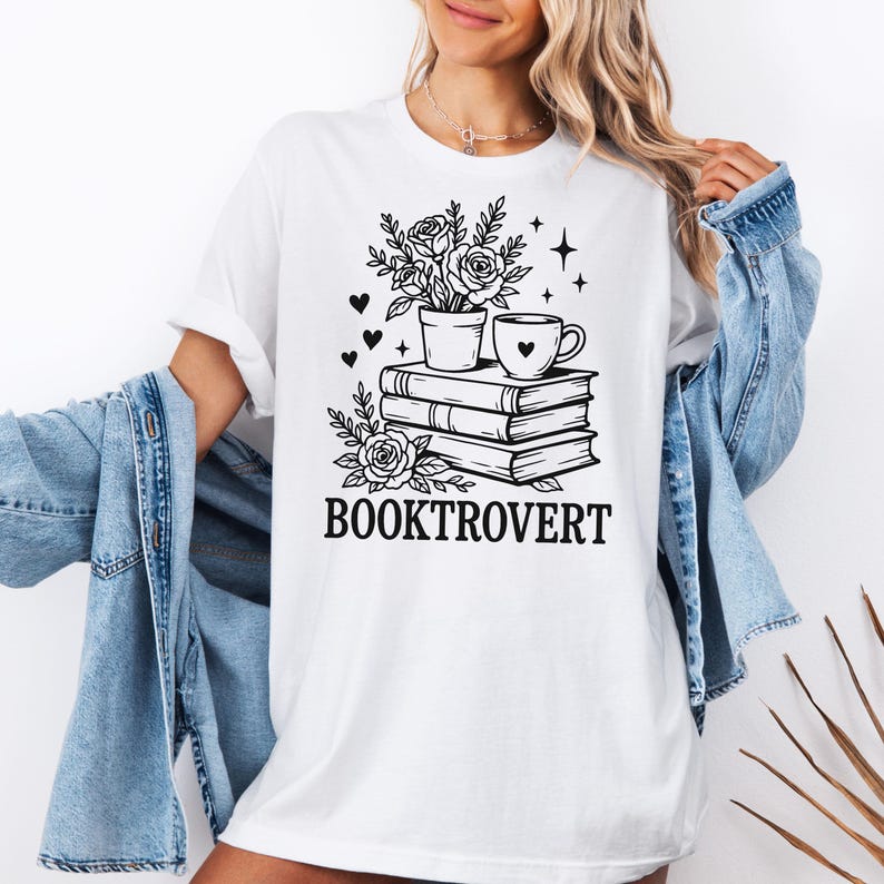 May include: White t-shirt with a black graphic of a potted plant, a coffee cup with a heart, and a stack of books. The word "BOOKTROVERT" is printed below the graphic. The shirt is made of a soft material.
