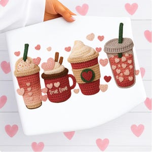 May include: A white surface displays four crocheted coffee cups and mugs in shades of red, pink, and beige, adorned with heart-shaped embellishments. One mug reads "true love." The cups have green and brown accents.