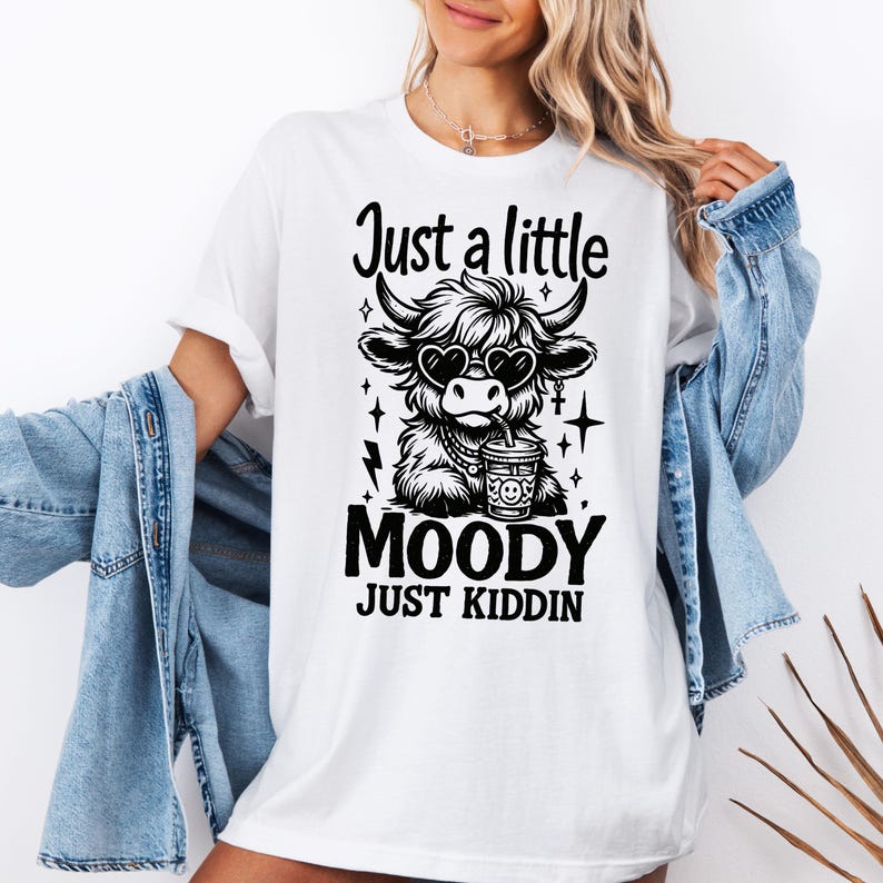 K&ouml;nnte beinhalten: Wei&szlig;es T-Shirt mit einer schwarzen Grafik einer Kuh mit herzf&ouml;rmiger Sonnenbrille und einem Getr&auml;nk. Der Text lautet "Just a little Moody Just Kiddin".
